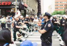 A police officer oversees a marching band parade in a busy urban street setting.