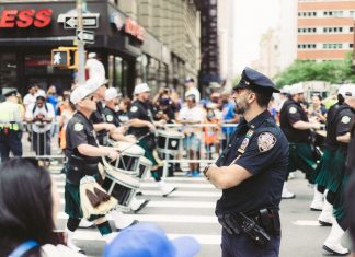 A police officer oversees a marching band parade in a busy urban street setting.