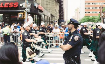 A police officer oversees a marching band parade in a busy urban street setting.