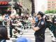 A police officer oversees a marching band parade in a busy urban street setting.