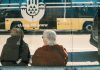 Jersey City Seniors to Get Microtransit Boost from Gambling Revenue Two individuals at a bus stop with a yellow bus reflection creating an urban scene.