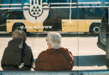 Jersey City Seniors to Get Microtransit Boost from Gambling Revenue Two individuals at a bus stop with a yellow bus reflection creating an urban scene.