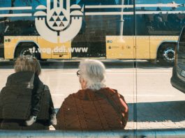 Two individuals at a bus stop with a yellow bus reflection creating an urban scene.