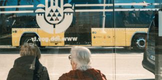 Two individuals at a bus stop with a yellow bus reflection creating an urban scene.