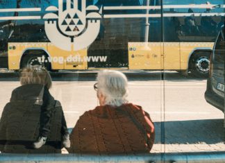 Two individuals at a bus stop with a yellow bus reflection creating an urban scene.