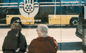 Two individuals at a bus stop with a yellow bus reflection creating an urban scene.
