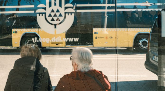 Jersey City Seniors to Get Microtransit Boost from Gambling Revenue Two individuals at a bus stop with a yellow bus reflection creating an urban scene.