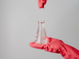 Laboratory scene showing red-gloved hands holding a flask with clear liquid.