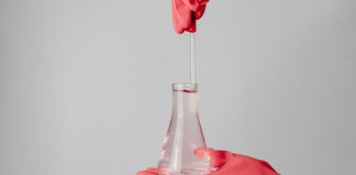 Laboratory scene showing red-gloved hands holding a flask with clear liquid.