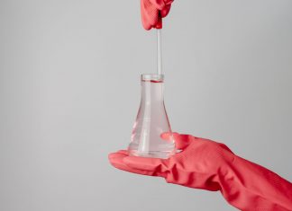 Laboratory scene showing red-gloved hands holding a flask with clear liquid.