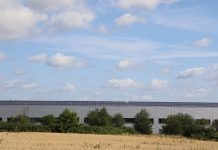 A countryside warehouse with solar panels and fields under a blue sky with clouds.