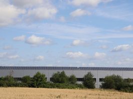 A countryside warehouse with solar panels and fields under a blue sky with clouds.
