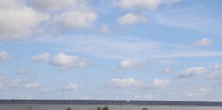 A countryside warehouse with solar panels and fields under a blue sky with clouds.