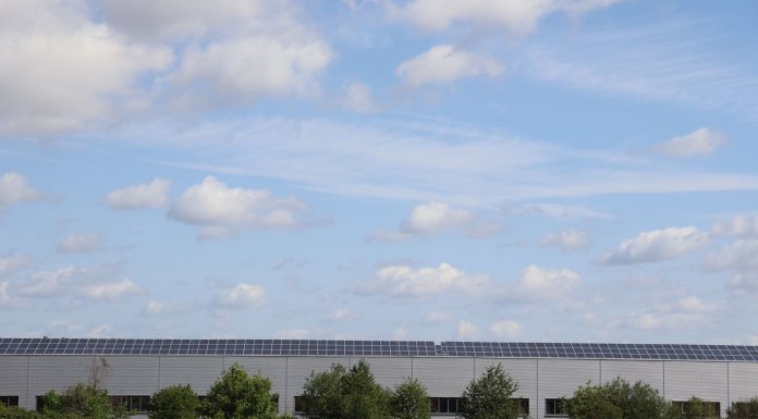 A countryside warehouse with solar panels and fields under a blue sky with clouds.