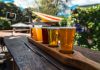 A vibrant beer flight displayed on a rustic wood table outdoors during a sunny day.