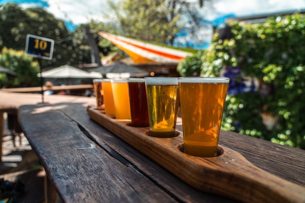 A vibrant beer flight displayed on a rustic wood table outdoors during a sunny day.
