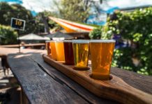A vibrant beer flight displayed on a rustic wood table outdoors during a sunny day.