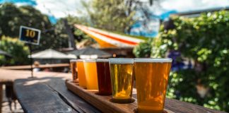 A vibrant beer flight displayed on a rustic wood table outdoors during a sunny day.