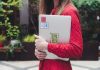 A young woman in a red polka dot shirt holds a laptop with stickers outdoors.