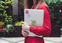 A young woman in a red polka dot shirt holds a laptop with stickers outdoors.
