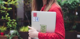 A young woman in a red polka dot shirt holds a laptop with stickers outdoors.