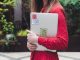 A young woman in a red polka dot shirt holds a laptop with stickers outdoors.