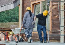 Two construction workers wearing hard hats work at an outdoor site, one with a wheelbarrow.