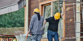 Two construction workers wearing hard hats work at an outdoor site, one with a wheelbarrow.