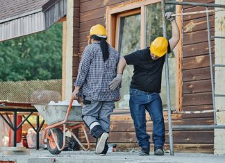 Two construction workers wearing hard hats work at an outdoor site, one with a wheelbarrow.