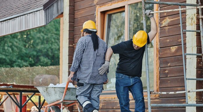 Two construction workers wearing hard hats work at an outdoor site, one with a wheelbarrow.