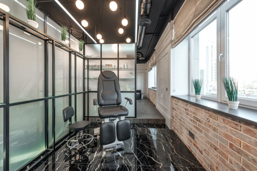 Black leather pedicure chair placed between glass door and window in modern beauty salon with decorated plants and glowing lamps
