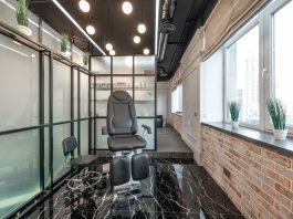 Black leather pedicure chair placed between glass door and window in modern beauty salon with decorated plants and glowing lamps