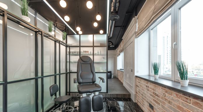 Black leather pedicure chair placed between glass door and window in modern beauty salon with decorated plants and glowing lamps