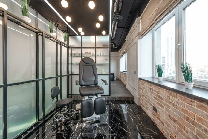 Black leather pedicure chair placed between glass door and window in modern beauty salon with decorated plants and glowing lamps