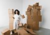 Young female with curly hair between piles and flying cardboard containers after moving into house on light background