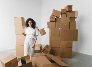 Young female with curly hair between piles and flying cardboard containers after moving into house on light background