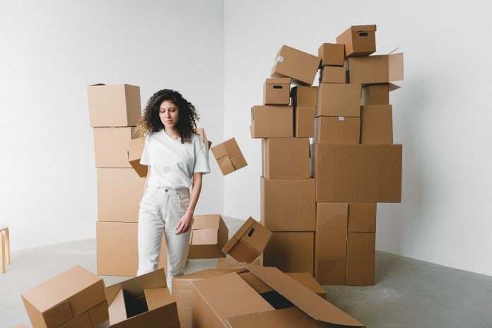 Young female with curly hair between piles and flying cardboard containers after moving into house on light background