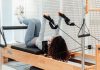 A woman wearing a gray tracksuit exercises on a Pilates reformer in a modern studio setting.