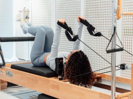 A woman wearing a gray tracksuit exercises on a Pilates reformer in a modern studio setting.