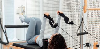 A woman wearing a gray tracksuit exercises on a Pilates reformer in a modern studio setting.