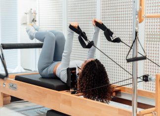 A woman wearing a gray tracksuit exercises on a Pilates reformer in a modern studio setting.