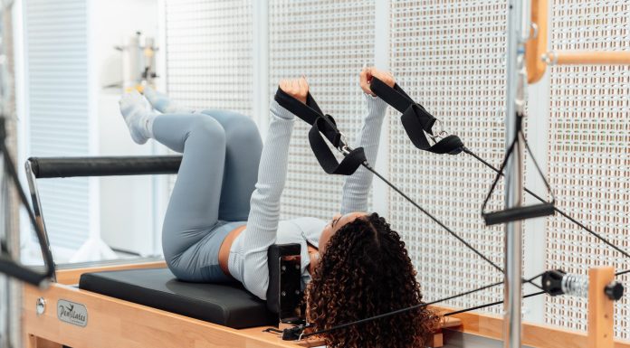 A woman wearing a gray tracksuit exercises on a Pilates reformer in a modern studio setting.
