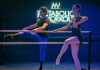 Two ballet dancers practice at a barre under a neon sign in an indoor studio.