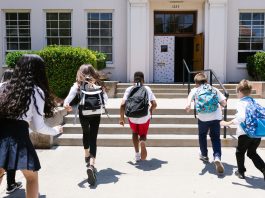 Group of students with backpacks running into school, captured from behind, showing excitement.
