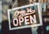 A welcoming 'Come in, We Are Open' sign hanging on a glass window of an urban shop.