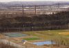Aerial view of multi-sport fields in urban park with city skyline and bridge in background.