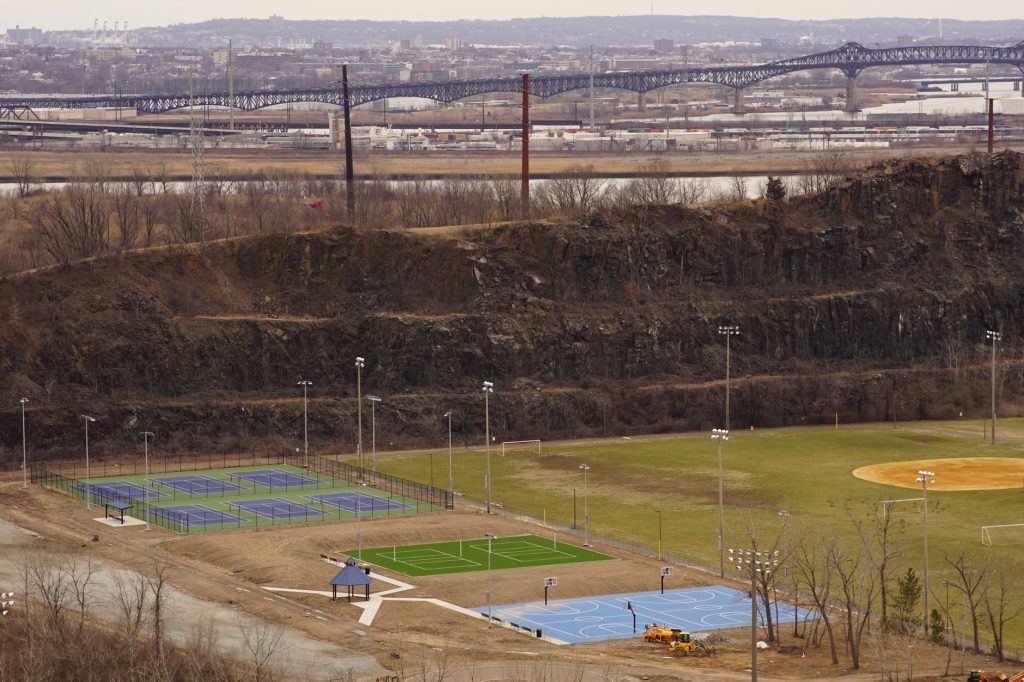 Aerial view of multi-sport fields in urban park with city skyline and bridge in background.
