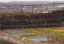 Aerial view of multi-sport fields in urban park with city skyline and bridge in background.