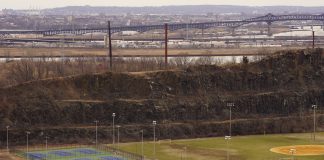 Aerial view of multi-sport fields in urban park with city skyline and bridge in background.