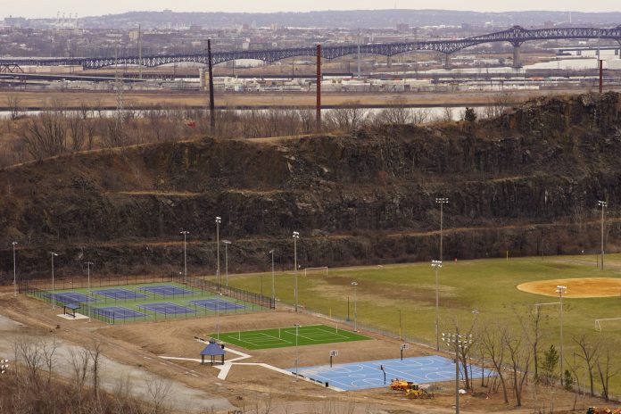 Aerial view of multi-sport fields in urban park with city skyline and bridge in background.